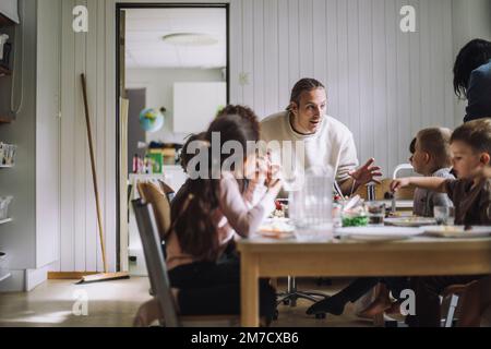 Männlicher Lehrer, der mit Schülern spricht, frühstückt am Tisch in der Kindertagesstätte Stockfoto