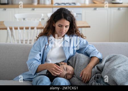 Eine gute fürsorgliche Frau sitzt auf dem Sofa und umarmt ein Mädchen, das während der Krankheit auf Knien in eine warme Decke gewickelt liegt Stockfoto