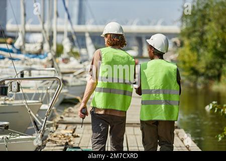 Rückansicht von zwei Arbeitern, die Schutzhelme tragen, während sie auf dem Pier in den Yachtdocks spazieren, Kopierbereich Stockfoto