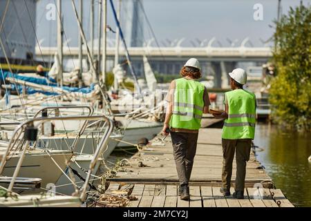 Volle Rückansicht von zwei Arbeitern mit Schutzhelm, während sie auf dem Pier spazieren, Kopierbereich Stockfoto
