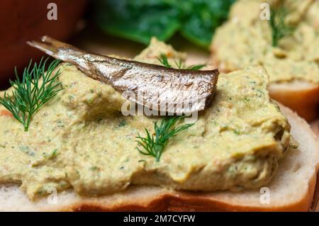 Sandwich mit Pastete und Sprotten. Sprottenpastete mit Spinat und Pfeffer. Sprotte in Öl, dänische Küche. Hintergrund zu Lebensmittelrezepten. Sandwich mit geräucherter Sprotte. Stockfoto