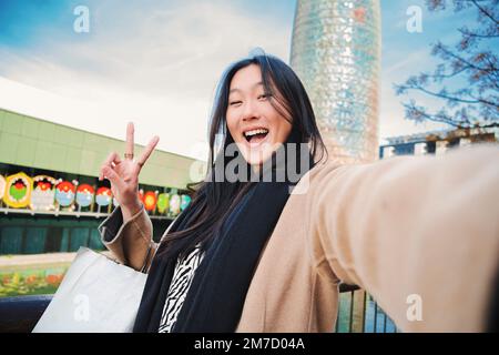 Glückliche junge asiatische Frau, die ein Selfie-Foto mit einem Smartphone macht. Selbstporträt einer lächelnden chinesin, die nach dem Einkaufen im Freien das Friedenszeichen trägt. Hochwertiges Foto Stockfoto