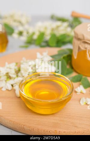 Süßes Honigglas umgab die Akazienblüten des Frühlings. Honig fließt aus einem Löffel in ein Glas. Gläser von klarem frischen Akazienhonig auf Holzgrund. Stockfoto