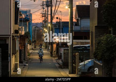 Akashi, Japan - 29. Dezember 2022: Radfahrer fahren bei Sonnenuntergang durch eine enge Wohngasse Stockfoto