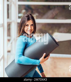 Ein Mädchen mit Matte, bereit für einen Yoga-Kurs. Sie sieht lächelnd aus und trägt blaue Leggings. Stockfoto