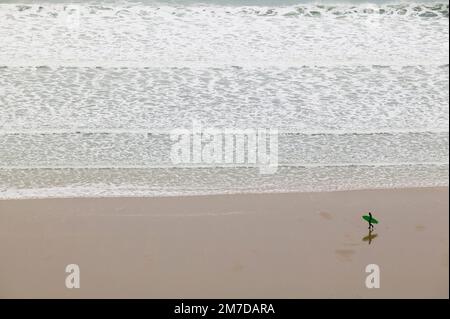 Ein einsamer Surfer spaziert am langen Sandstrand von Wollacombe in Devon, Großbritannien. Die Silhouette spiegelt sich im nassen Sand wider, während er an den Reihen oder Wellen vorbeigeht, die in einem Winterschwall in den Strand kommen. Stockfoto