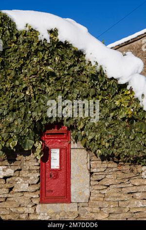 Ein tiefes roter traditionellen Briefkasten inmitten einer Trockensteinmauer in den Cotswolds, Gloucestershire Uk, Efeu und mit einer Schicht aus Schnee an der Spitze in diesem Winter Landschaft Szene abgedeckt. Stockfoto