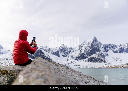 Mann, der im Winter mit Smartphone die gefrorene Landschaft fotografiert, Flakstadpollen, Nordland County, Lofoten-Inseln, Norwegen Stockfoto