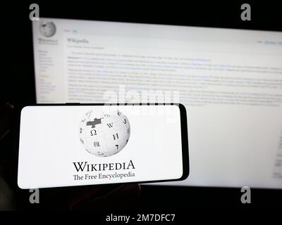 Person, die ein Smartphone mit dem Logo der Online-Enzyklopädie Wikipedia auf dem Bildschirm vor der Website hält. Konzentrieren Sie sich auf das Display des Telefons. Stockfoto