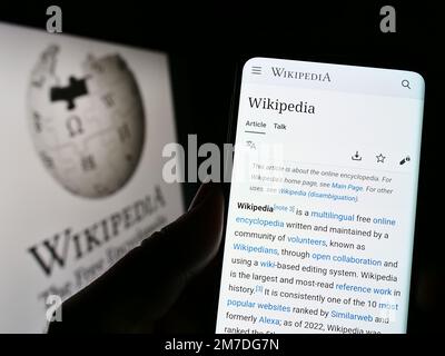 Person, die ein Mobiltelefon mit einer Webseite der Online-Enzyklopädie Wikipedia auf dem Bildschirm vor dem Logo hält. Konzentrieren Sie sich auf die Mitte des Telefondisplays. Stockfoto