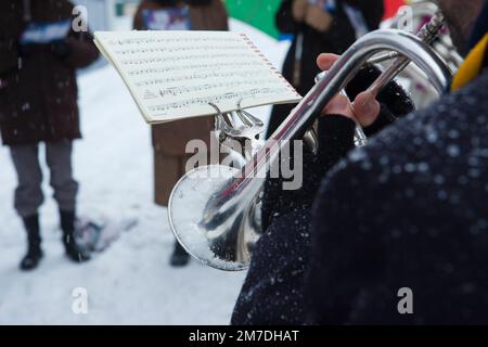 Die Band der Heilsarmee spielt Weihnachtslieder auf einem cotswold-Marktplatz, während Sänger dabei sind, im Winter Schnee zu weihnachten. Stockfoto