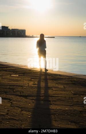 Silhouette "Frau allein", Rückansicht einer jungen Frau, die allein im hellen Sonnenlicht steht und auf das Meer blickt. Stockfoto