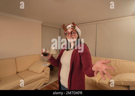 Eine wunderschöne junge hispanikerin, verkleidet mit Rentiergläsern und einem weihnachtshut, die in die Kamera schaut und mit einem Glas Rotwein anstoßen Stockfoto