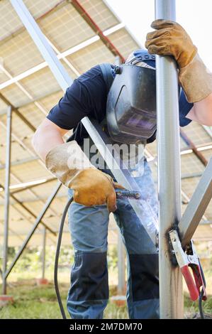 Schweißer-Metallbalken mit Schweißgerät zur Installation von Solarpaneelen. Konzept der erneuerbaren und ökologischen Energie. Vorstellung von umweltverträglich. Arbeiter im Schweißschutzhelm in Arbeitskleidung Stockfoto