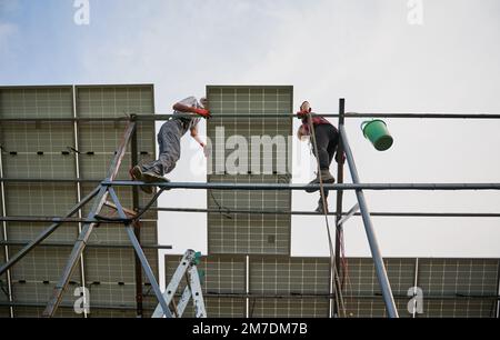 Blick von unten auf zwei Solartechniker, die auf einer Metallschiene stehen und Photovoltaik-Solarpaneele montieren. Männliche Arbeiter, die Solarmodule unter weißem blauen Himmel installieren. Konzept der nachhaltigen Energie. Stockfoto