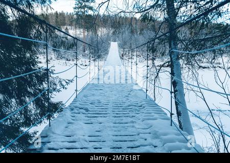 Wunderschöne, an einem Seil hängende Holzbrücke über den Fluss. Winterlandschaft, schneebedeckter Wald. Perspektive, Symmetrie Stockfoto