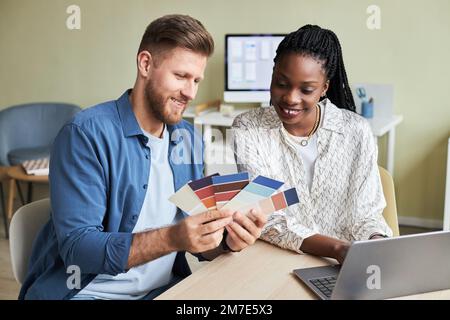 Porträt von zwei lächelnden jungen Designern, die Farbmuster betrachten, während sie im Büro an kreativen Projekten arbeiten Stockfoto
