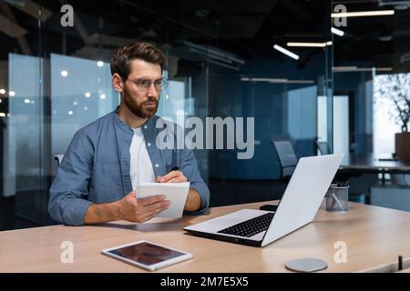 Ein seriöser Geschäftsmann schreibt Informationen auf, während er im Büro sitzt und online studiert. Stockfoto