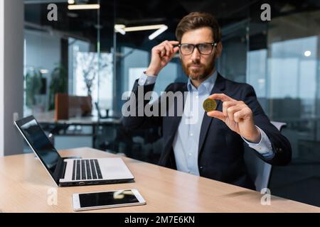 Seriöser und fokussierter Geschäftsmann, der in die Kamera schaut, reifer erfolgreicher Investor, der eine Kryptowährung mit Bitmünzen hält und auf ihn zeigt, bärtiger Mann, der in einem modernen Büro arbeitet. Stockfoto