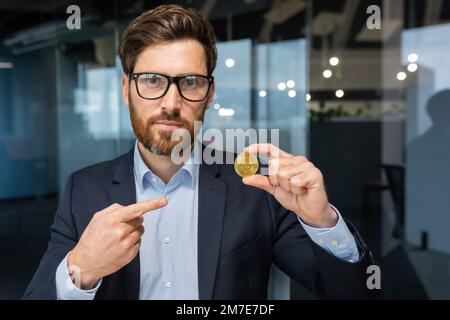 Seriöser und fokussierter Geschäftsmann, der in die Kamera schaut, reifer erfolgreicher Investor, der eine Kryptowährung mit Bitmünzen hält und auf ihn zeigt, bärtiger Mann, der in einem modernen Büro arbeitet. Stockfoto