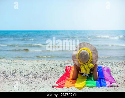 Eine Frau, die auf dem Rücken am Meer auf einer stolzen Flagge sitzt. Sie trägt einen Strohhut und einen gelben Schal. lgtb. Begriff von Vielfalt und Toleranz. Stockfoto