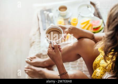 Blick von oben auf eine Frau, die im Hotelbett frühstückt und ihre Tasse Kaffee hält. Konzept von Sommerurlaub und Entspannung. Stockfoto