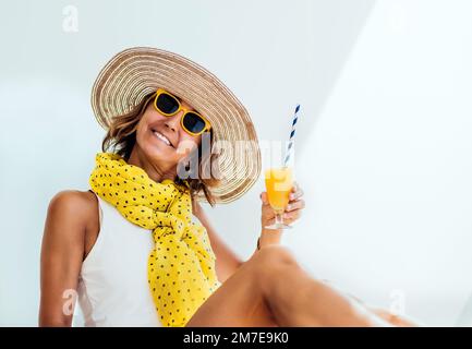 Porträt einer Frau mittleren Alters, die in die Kamera schaut, einen Hut und eine gelbe Brille trägt und einen Orangensaft in der Hand hält. Urlaubskonzept Stockfoto