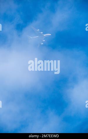 Ein Düsenflugzeug fliegt über den Kopf durch eine Lichtwolke, die hinter der weißen Schicht vor einem blauen Himmel verschwindet. Stockfoto