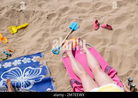 Weibliche Füße mit Beinen, die auf einem Handtuch liegen, umgeben von Urlaubsgegenständen, Eimer, Spaten und Dosen mit Getränken, während wir auf einen Sandstrand blicken. Stockfoto