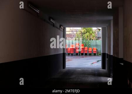 Stapel roter Kunststoffstühle, die im Garten gelagert werden und durch einen langen Tunnel unter Gebäuden sichtbar sind. Stockfoto