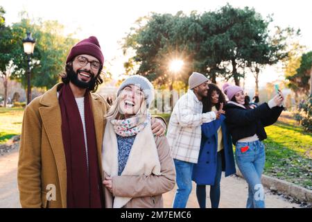 Ein junges, nettes Paar, das lächelt und lacht, in die Kamera schaut, im Hintergrund drei Freunde, die ein Selfie-Foto mit einem Handy machen, einen Schal, Hüte, Mäntel und Winter- oder Herbstkleidung tragen. Lifestyle-Konzept. Hochwertiges Foto Stockfoto