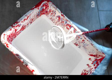 Ein offener Kunststoffeimer mit weißer Farbe und ein Rührer, der auf dem Boden steht. Stockfoto