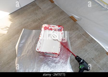 Ein offener Kunststoffeimer mit weißer Farbe und ein Rührer, der auf dem Boden steht. Stockfoto