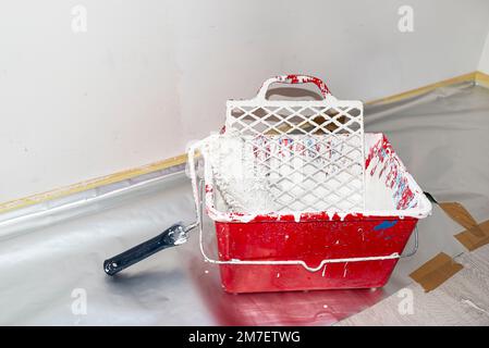 Ein offener Kunststoffeimer mit weißer Farbe und eine Malerwalze, die auf dem Boden steht. Stockfoto