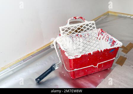 Ein offener Kunststoffeimer mit weißer Farbe und eine Malerwalze, die auf dem Boden steht. Stockfoto