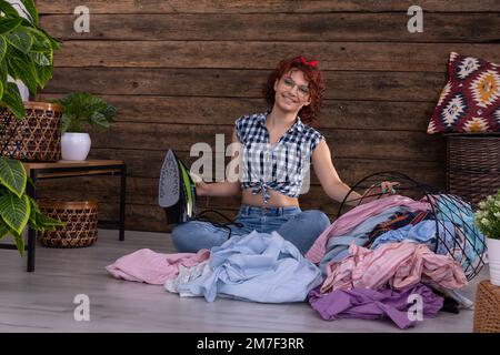 Das Mädchen sitzt auf dem Boden und hält ein Bügeleisen in der Hand. Ein Lächeln auf ihrem Gesicht. Stockfoto