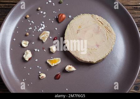 Französische Foie Gras serviert mit Nüssen, Pfeffer, Salz und getrockneten Früchten auf einem braunen Teller, Nahaufnahme aus der Vogelperspektive Stockfoto