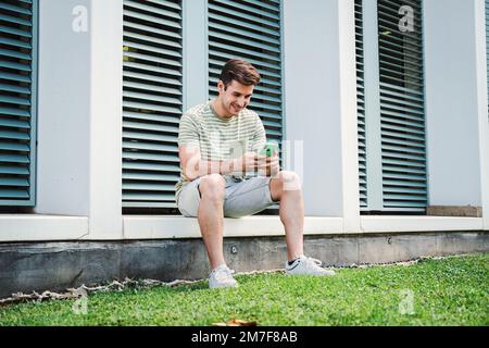 Ein junger Kerl, der Spaß hat, ein Spiel mit einem Handy zu spielen, das nach dem Unterricht auf dem Universitätscampus sitzt. Ein Schüler im Teenageralter, der mit einem Smartphone über eine Social-Media-App lächelt. Hochwertiges Foto Stockfoto