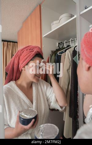Wunderschöne hispanische Frau in einem weißen Bademantel und einem roten Handtuch im Haar, die Make-up aufträgt, mit der Hand auf dem Gesicht vor dem Spiegel auf dem Schrank Stockfoto