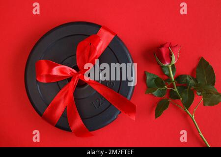 Gewichtsteller mit Kurzhantel und roter Rose. Liebesgeschenk zum Valentinstag, Geburtstag, Jubiläum oder zur Hochzeit. Fitnessstudio Workout flach liegend. Stockfoto