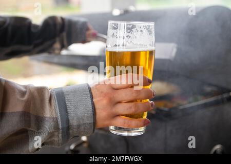 Weibliche Hand hält ein Glas Bier, während sie auf einem Grill kocht. Stockfoto