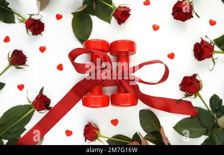 Die Kurzhanteln im Fitnessstudio sind als Geschenk zum Valentinstag, zum Jahrestag oder zur Hochzeit in ein Band eingewickelt. Gesunde Fitness-Workout flach legt mit roten Rosen und Herzen. Stockfoto