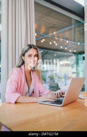 Junge Millennials-Führungskraft in einem pinkfarbenen Blazer, die ihren Computer an einem offenen Arbeitsplatz verwendet. Stockfoto
