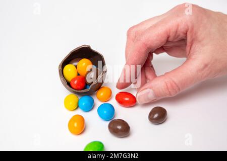 Eine Hand, die eine Süßigkeit aus einem zerbrochenen Schokoladenei nimmt, das voll von bunten, runden Süßigkeiten auf weißem Hintergrund mit bunten Süßigkeiten ist. Schokolade Stockfoto