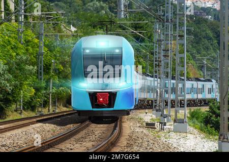 Der blaue Vorstadtzug dreht sich aus der Ferne mit eingeschalteten Scheinwerfern auf dem Weg zwischen den grünen Bäumen Stockfoto