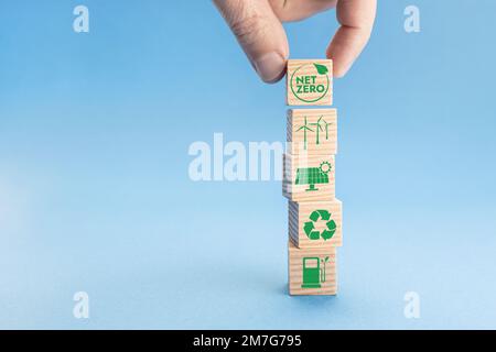 Netto-Null- und CO2-neutrales Konzept. Hand legt Holzblöcke mit dem Net Zero-Symbol über andere mit grünen Energiesymbolen. Blauer Hintergrund. Speicherplatz kopieren Stockfoto