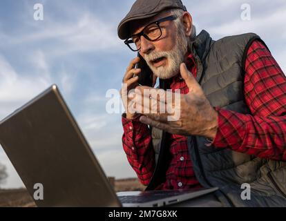 Ein seriöser älterer Bauer, der telefoniert und auf dem Feld mit offenem Mund in einen Laptop schaut Stockfoto