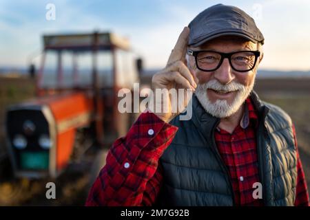 Porträt eines glücklichen, reifen Landwirts, der im Herbst vor dem Traktor auf dem Feld lächelt und grüßt Stockfoto