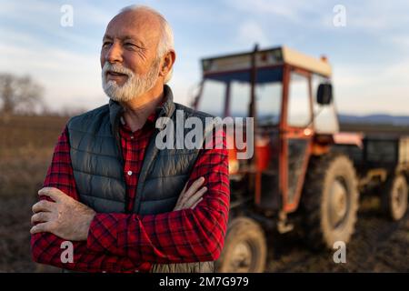 Porträt eines älteren Landwirts mit weißem Haar und Bart, der im Spätherbst vor dem Traktor auf dem Feld steht Stockfoto