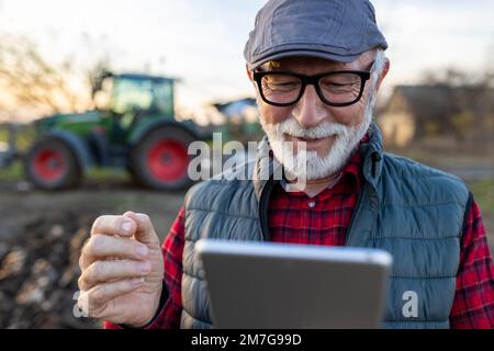 Lächelnder, reifer Landwirt, der auf dem Feld vor dem Traktor auf ein Tablet schaut Stockfoto